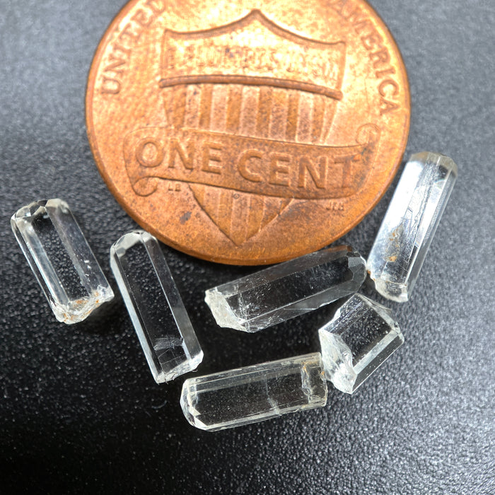 Top view of 6 pieces of Burmese phenakite hexagonal barrel stones on black background next to one cent piece for size. available at sanctuaryofstones.com