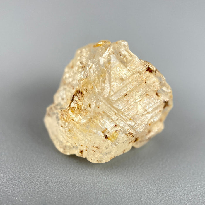 Etched rough Nigerian phenakite crystal with orange tint on grey background. available at sanctuaryofstones.com